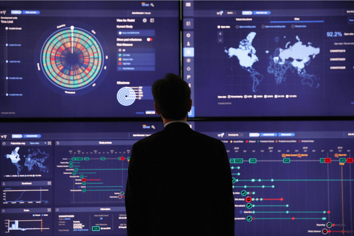 An analyst standing in front of the six-screen SENSE Bridge control center, with study overview, world map, and process timelines spanning the wall